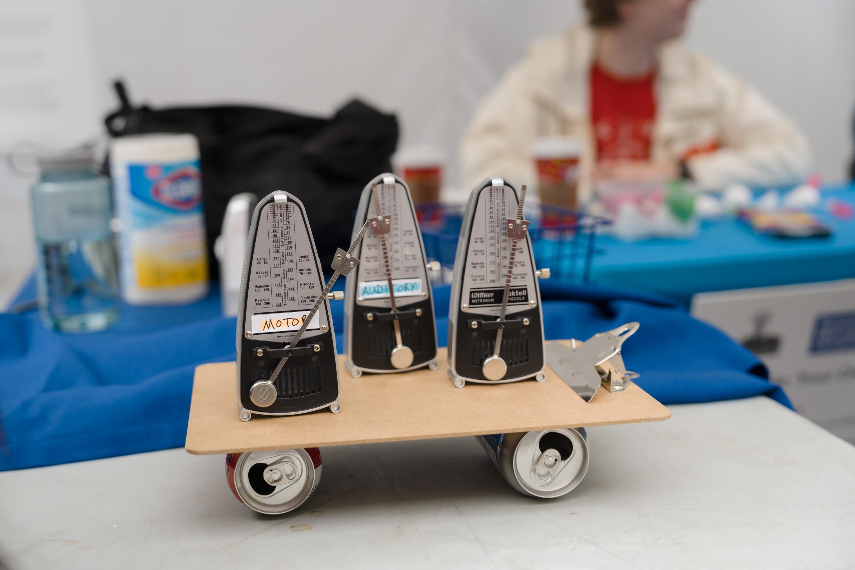 Three tiny metronomes with two labelled "motor" and "auditory" on a clipboard were balanced by two soda cans at the Mind Games Booth.
